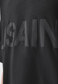 Țesătură neagră cu litere mari, glossy și majuscule care scriu o parte din numele brandului "ALSAIN" pe un articol vestimentar.