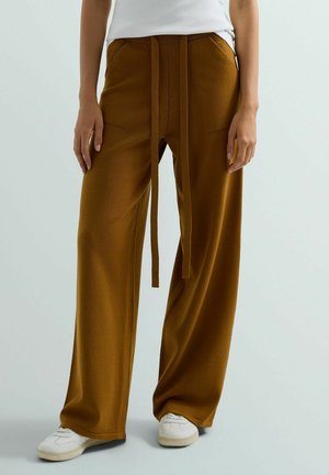 Donna che indossa pantaloni marroni a gamba larga con tasche frontali e sneakers bianche, in piedi su uno sfondo grigio chiaro.