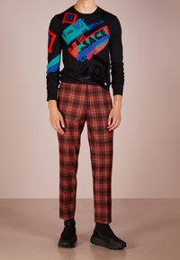 Zwarte sweater met kleurrijke tekst en geometrische ontwerpen, gecombineerd met rood-groene geruite broek. Compleet met zwarte sportschoenen.