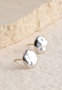 Silver stud earrings with an organic, irregular shape, featuring a polished surface and subtle texture against a soft background.