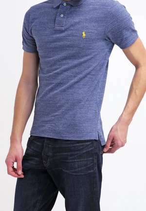 Mężczyzna ubrany w dopasowaną niebieską koszulkę polo z żółtym logo i ciemnoniebieskie dżinsy, stojący z jedną ręką blisko kieszeni na białym tle.