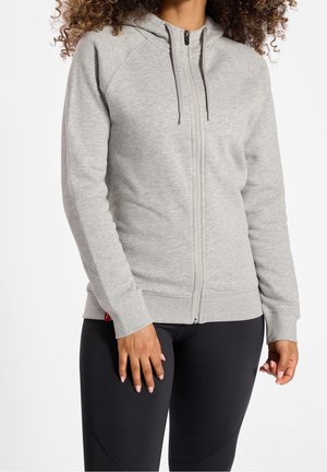 Femme portant un sweat à capuche gris clair zippé avec cordons et un pantalon noir ajusté, debout, les bras détendus le long du corps.