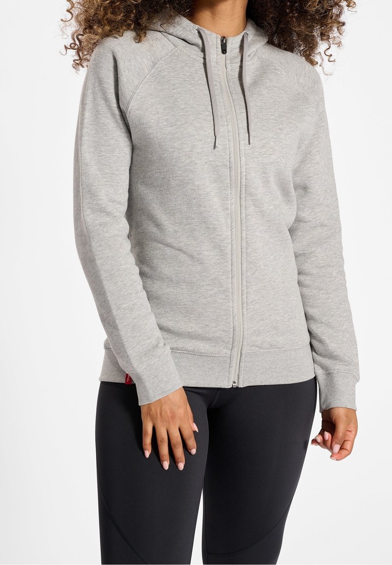 Femme portant un sweat à capuche gris clair zippé avec cordons et un pantalon noir ajusté, debout, les bras détendus le long du corps.