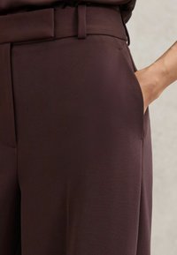 Tumši brūnas piegrieztas bikses ar gludu faktūru, kurās ir sānu kabata un jostas cilpa viduklī. Tīras līnijas un klasiskas dizains.