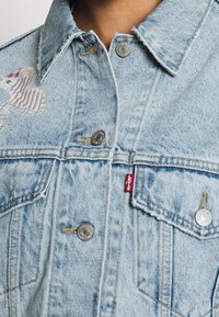 Giacca di jeans azzurro chiaro con bottoni argentati, bordi del colletto sfilacciati e un dettaglio ricamato colorato con un motivo a righe.