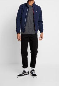 Marineblauer Bomberjacke mit roten Reißverschlussdetails, grauem T-Shirt, schwarzen hochgekrempelten Hosen und schwarzen Vans Sneakers mit weißen Akzenten.