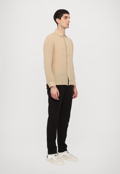 Camicia button-up a maniche lunghe beige chiaro in un tessuto testurizzato, abbinata a pantaloni neri e sneakers bianche, in piedi contro uno sfondo bianco.