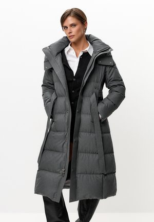 Mujer con abrigo de invierno largo gris acolchado con cuello grande, superpuesto sobre un cárdigan negro y camisa blanca, manos en los bolsillos, fondo neutro.