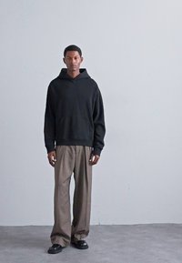 Homme debout contre un mur gris uni, portant un sweat à capuche noir, un pantalon marron ample et des chaussures noires, faisant face à l'avant avec une expression neutre.