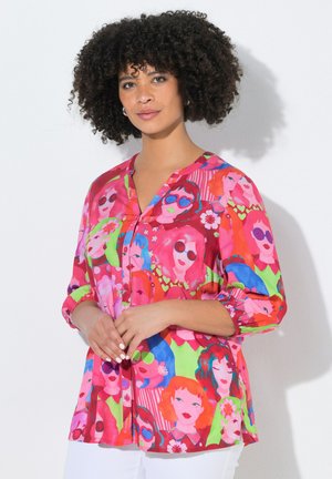 Frau mit lockigem Haar, die eine bunte Bluse trägt, bedeckt mit hellen, stilisierten Gesichtern mit Sonnenbrillen und Blumen, stehend vor einem weißen Hintergrund.