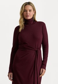 Donna bionda che indossa un vestito lungo a maniche con collo alto color bordeaux, con un nodo laterale, in piedi contro uno sfondo chiaro e semplice.