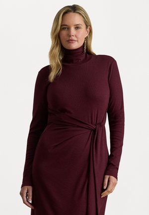 Blond naine, kes kannab burgundi värvi pikkade varrukatega kõrge kaelusega kleiti, millel on külgõhk, seistes lihtsa heledat tooni tausta vastu.