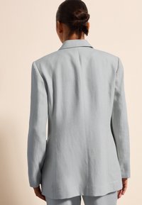 Blazer de lino azul claro con un diseño estructurado, solapa de muesca y silueta alargada. Textura suave con una abertura sencilla en la parte de atrás.