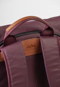 Zaino color amaranto con una texture liscia, caratterizzato da una tracolla in pelle marrone e un'etichetta in pelle con marchio. Il design è elegante e moderno.