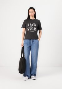 Must musta T-särk, millel on tekst "ROCK STAR", sobitatud helesiniste laia jala teksadega. Mudel hoiab musta kotti ja kannab valgeid tosse.