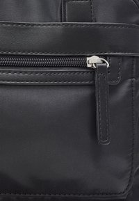Section d'un sac en tissu noir avec une poche zippée, présentant des accents en cuir lisse et des coutures détaillées. Tirette de fermeture éclair métallique et étiquette visibles.