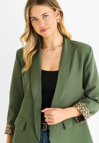 Blazer verde oliva con apertura frontale, tasche frontali e polsini con stampa leopardata. Abbinato a un top nero a costine e accessori in tono oro.