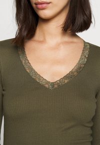 Olivgrünes, geripptes Langarmoberteil mit einem floralen Spitzenbesatz um den V-Ausschnitt, der Textur und zarte Details betont.