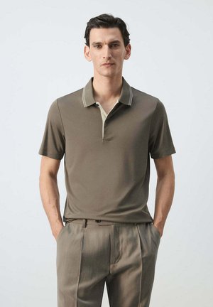 Homme debout devant un fond uni, portant un polo taupe à manches courtes et un pantalon plissé assorti, les mains dans les poches.