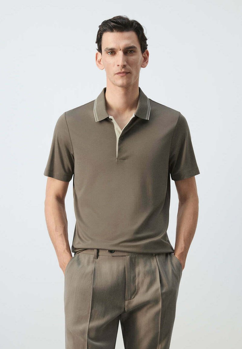 Man standing against a plain background, wearing a taupe short-sleeve polo shirt and matching pleated trousers, hands in pockets.