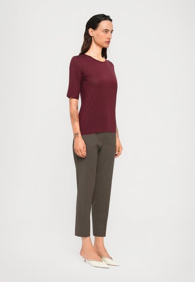 Woman standing sideways wearing a burgundy short-sleeve top, gray cropped pants, white pointed-toe heels, and visible arm tattoos.