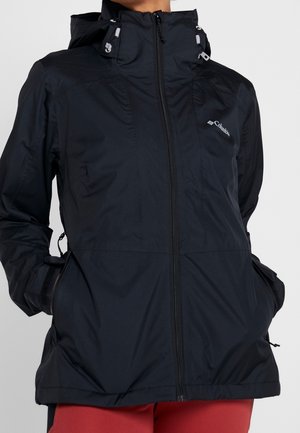 Person trägt eine schwarze Columbia-Kapuzenjacke mit Frontreißverschluss und den Händen in den Seitentaschen, kombiniert mit roten Hosen.