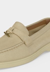 Mocassin en daim beige avec semelle en caoutchouc crème et détail d’une boucle métallique dorée sur le dessus, montrant la doublure intérieure marron.