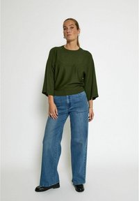 Un pull oversized vert à col rond associé à un pantalon en jean bleu à taille haute et jambes larges, porté avec des chaussures en cuir verni noires.