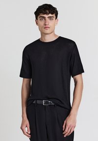 Giovane uomo con capelli ricci scuri che indossa una camicia nera a maniche corte con texture, cintura nera e pantaloni neri su uno sfondo neutro.