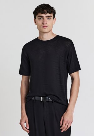 Jeune homme avec des cheveux bouclés et foncés, portant une chemise noire à manches courtes texturée, une ceinture noire et un pantalon noir devant un fond uni.