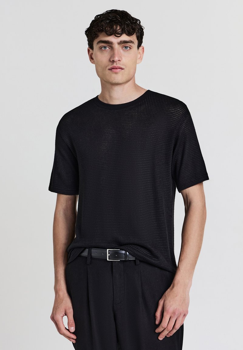 Giovane uomo con capelli ricci scuri che indossa una camicia nera a maniche corte con texture, cintura nera e pantaloni neri su uno sfondo neutro.