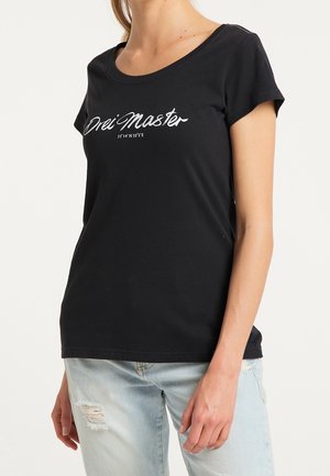 Frau trägt ein schwarzes T-Shirt mit dem Schriftzug "Drei Master" und Koordinaten, kombiniert mit hellblauen, zerrissenen Jeans.