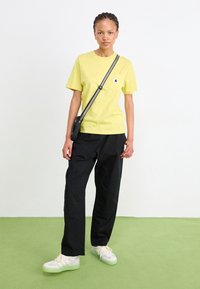 Jeune personne debout sur un sol vert portant un T-shirt jaune, un pantalon noir, des baskets blanches et un sac à bandoulière avec une sangle rayée.