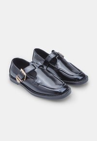 Zapatos de charol negros con puntera redonda, que presentan una correa con un hebilla en tono dorado y una suela de goma plana. Acabado suave.