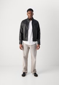 Jachetă din piele neagră cu detalii quilted orizontale, tricou alb dedesubt, pantaloni gri deschis și adidași negri cu accente albe.