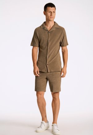 Hombre de pie con camisa marrón de manga corta con botones y pantalones cortos a juego, con zapatillas blancas sobre fondo blanco.