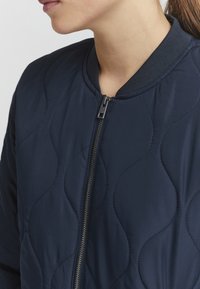 Marineblå quiltet bomberjakke med ribbet krave og en sølvfarvet lynlås. Stoffet har en glat tekstur med bølgeformede syningmønstre.