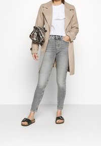 Un abrigo trench beige sobre una blusa blanca con volantes, combinado con unos jeans ajustados grises y sandalias negras de plantilla. Un bolso de mano a cuadros en la mano.