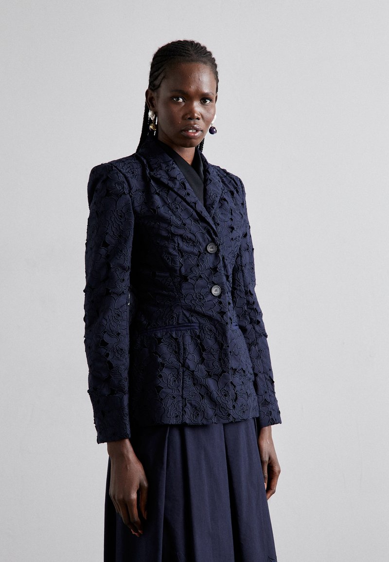 Navy blue lace blazer with floral pattern, two buttons, fitted design, and structured shoulders, worn over a matching skirt.