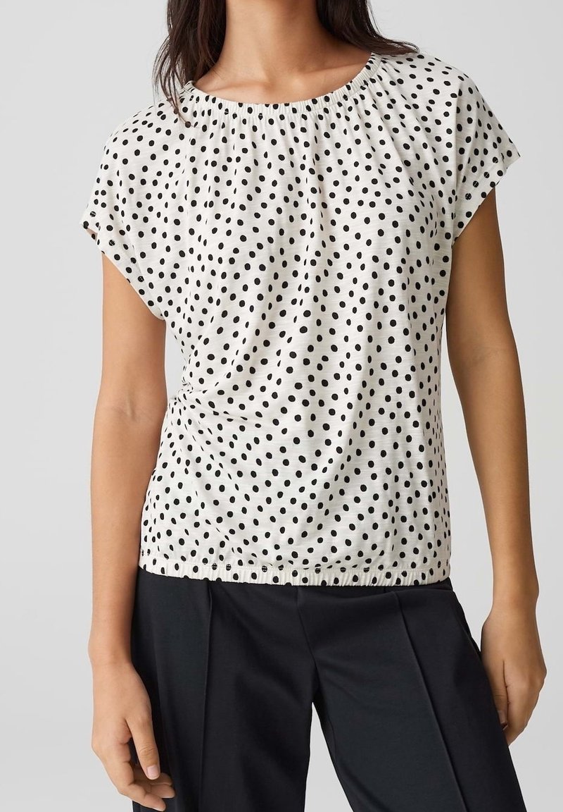 Femme portant un chemisier blanc à manches courtes avec des pois noirs et un pantalon noir, debout devant un fond uni.