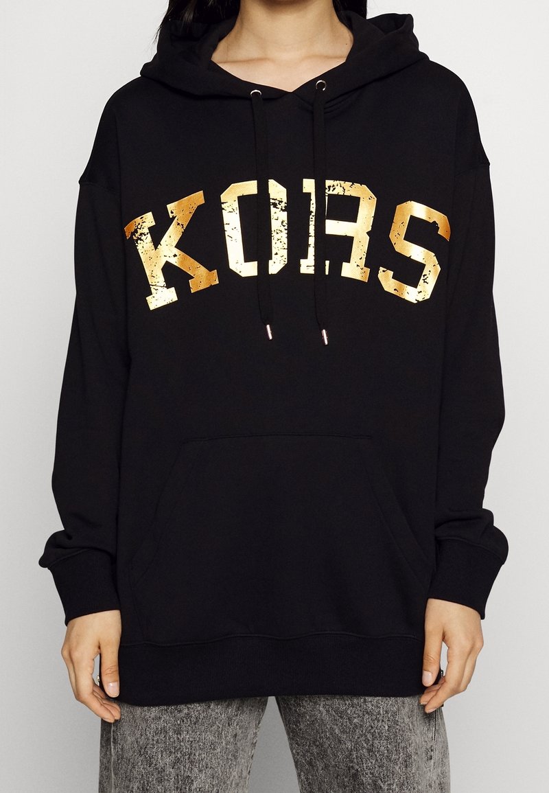 Personne portant un sweat à capuche noir avec texte métallique doré "KORS", cordons noirs et poche avant, associé à un jean gris délavé.
