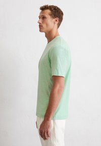 Hellgrünes T-Shirt mit kurzen Ärmeln aus weichem, strukturiertem Stoff mit Rundhalsausschnitt, präsentiert an einem Model, das seitlich vor einem neutralen Hintergrund steht.