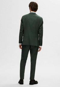 Zaļš teksturēts uzvalks ar šauru piegriezumu, ar vienu venti, noslēgtām lapelēm un saskaņotām biksēm. Pabeigts ar tumšām kurpēm.