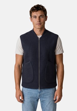 Giovane uomo con un gilet blu navy con cerniera sopra una maglietta bianca a maniche corte e jeans azzurri chiari, in piedi davanti a uno sfondo bianco semplice.