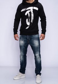 Czarna bluza z kapturem z białym logo w stylu pędzla, zestawiona z przetartymi niebieskimi dżinsami z detalami w postaci przetarć. Włożona z jasnymi trampkami.