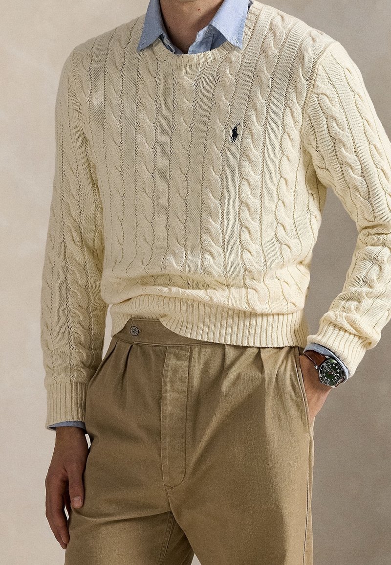 Personne portant un pull crème torsadé sur une chemise bleue à col, un pantalon beige plissé et une montre-bracelet avec un bracelet en cuir marron.