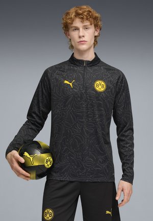 Sudadera deportiva negra con un patrón sutil, que presenta un logo de Puma amarillo y un escudo del Borussia Dortmund. Pantalones negros y un balón de fútbol negro y amarillo.