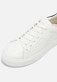 Baskets en cuir blanc avec lacets plats, bout arrondi et détails perforés sur le côté. Semelle en caoutchouc texturé pour une meilleure adhérence. Design minimaliste.