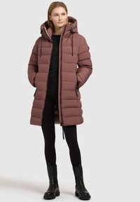 Piepūsts garais mētelis putekļu rozā krāsā ar kapuci, tajā ir auklas, rāvējslēdzēju un sānu kabatas; uzvilkts virs melnajām legingām un zābakiem.