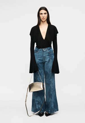 Ženska oblečena v črno bluzo z globokim V-izrezom in širokimi rokavi, modre kavbojke z visokim pasom in širokimi nogavicami, črne čevlje ter v roki zlato kovinsko ročno torbico.